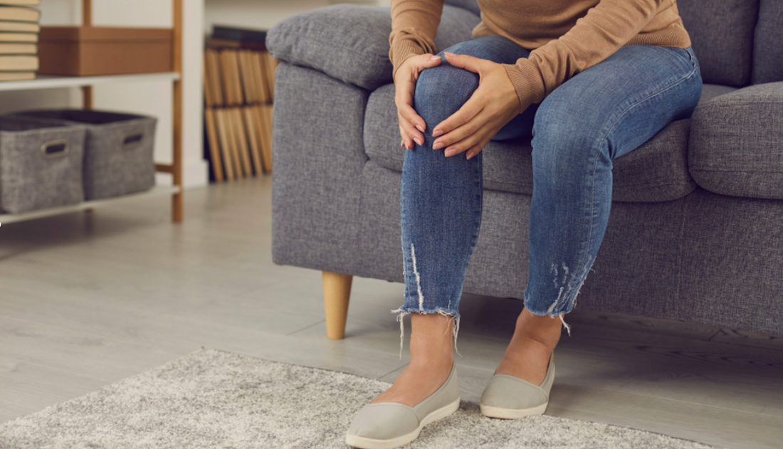Žena sedící na pohovce, držící si bolestivé koleno – symbol bolesti kloubů a revmatoidní artritidy.